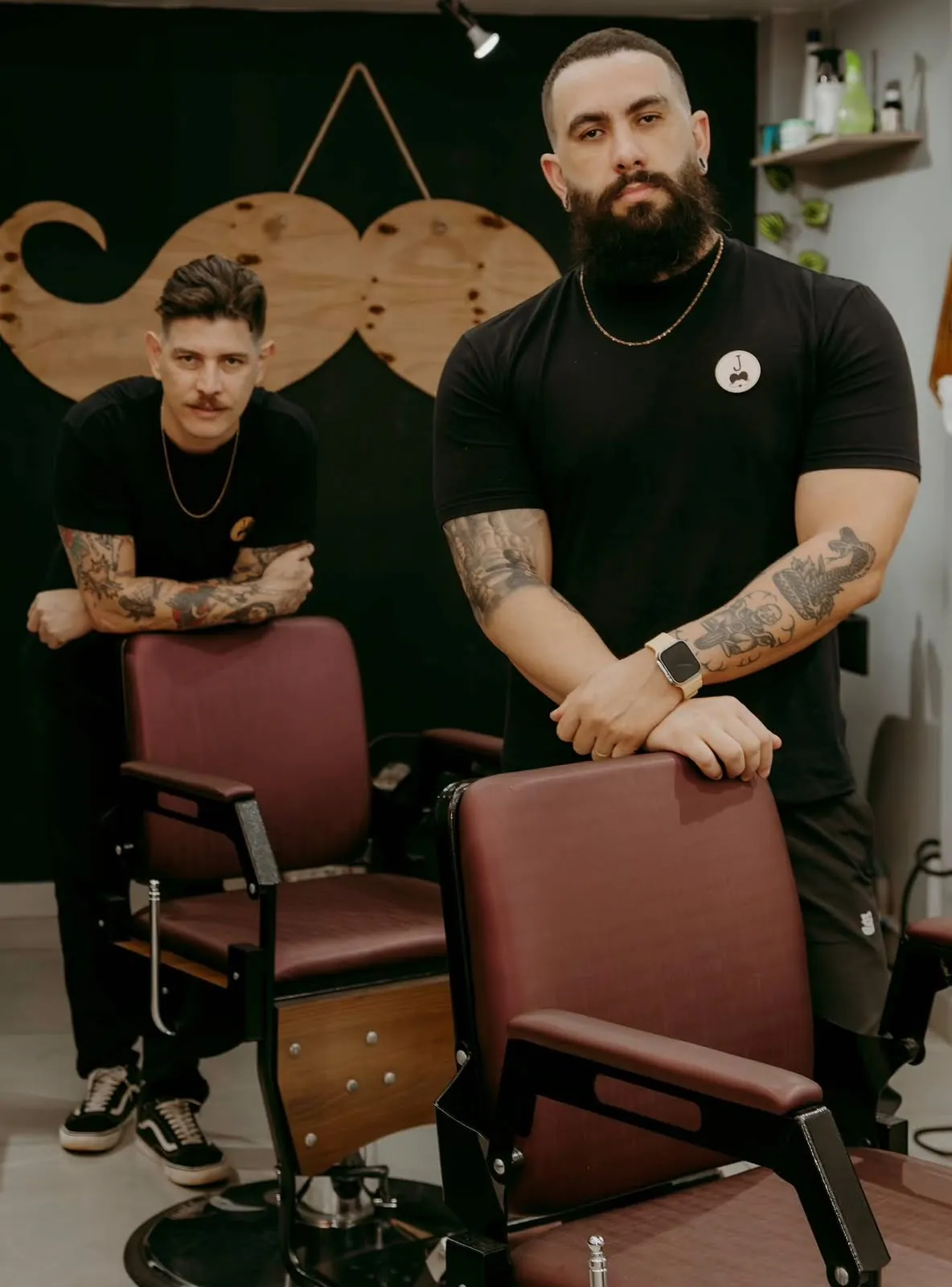Os dois barbeiros da Valete, de camiseta preta, apoiados nas cadeiras de barbeiro com o bigode de madeira na parede ao fundo