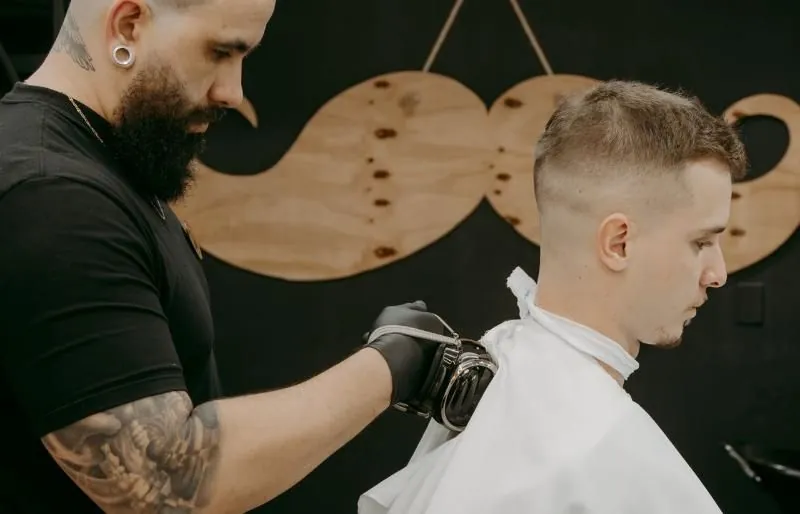 Barbeiro fazendo degradê com máquina, com o bigode de madeira ao fundo
